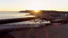 Drone pulls back from harbor and town of West Bay in Dorset as the sun sets over the distant hills.