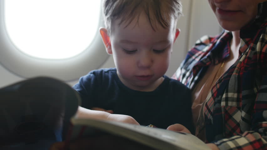 Plane is flying, the little kid is sitting at the mom on the pens, lookin children's magazine with pictures.