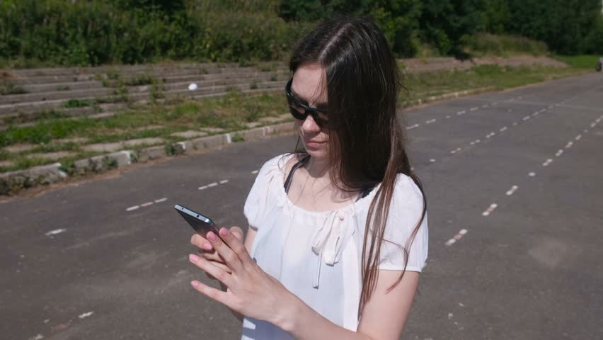 Young brunette girl walking in the stadium, dials a number on mobile phone and waiting for an answer.