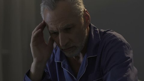 Elderly male sitting on bed in dark room, rubbing his temples, strong headache