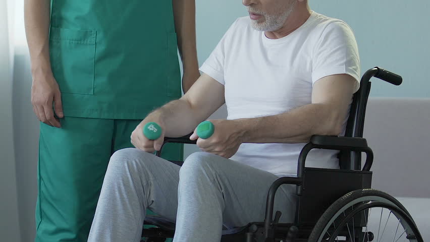 Aged man sitting in wheelchair, holding dumbbells, talking to nurse, recovery