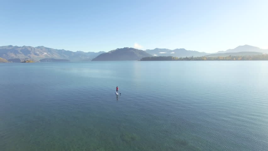 Aerial drone shot moving around athletic woman wearing pink shirt and paddle boarding towards the mountains on a beautiful clear lake surrounded by autumn trees