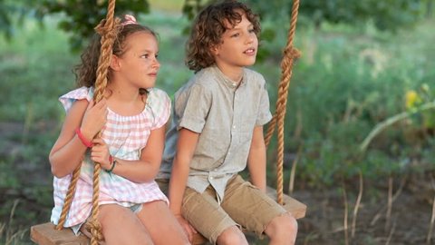 A pair of children on a swing. The boy kisses the girl on the cheek. The baby gives him a slap in the face. First love, summer holidays in the village