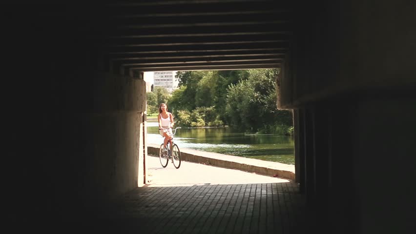  A tanned young woman in sunglasses in  white T-shirt with open shoulders  and in a gray skirt ride a vintage bicycle in park. A girl with a scar. A girl is riding a bicycle under a bridge.