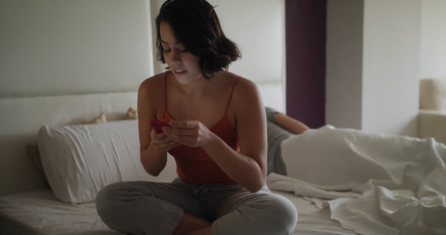 Young latino wife and husband in bed at home. Hispanic woman texting with smartphone and sending message to another man while her husband is still sleeping