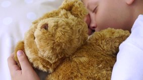 Closeup portrait of cute sleepy young white kid laying in his bed with cute brown teddy bear toy. Boy hugs happily his favorite soft toy. Real time 4k video footage.