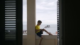 A young boy use his tablet for playing game while sits down on balcony at home at the sea background. Teenager playing video games on his smartphone outdoors in the summer, slow motion