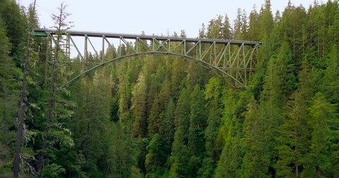 4K aerial shot flying under an old steel bridge in the deep forest of Washington state. The trees are tall and thick giving the bridge the appearance that its floating in the middle of nowhere.