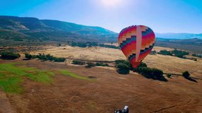 A colorful hot air balloon floating in the air slowly. The clip was recorded by a drone flying from a distance. The Mountains can be seen rising against the clear blue sky in the background.