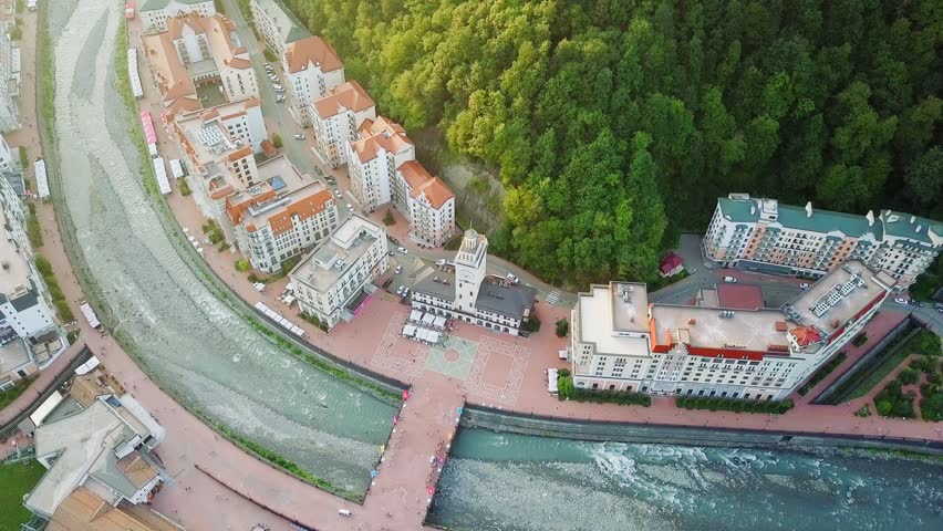 View from the air. Rosa Khutor alpine resort village in Krasnaya Polyana, Sochi, Russia, centered on the distinctive clock tower that serves as the symbolic heart and navigation landmark of the area. The tower stands at the convergence of the Mzymta River, which was straightened during the resort