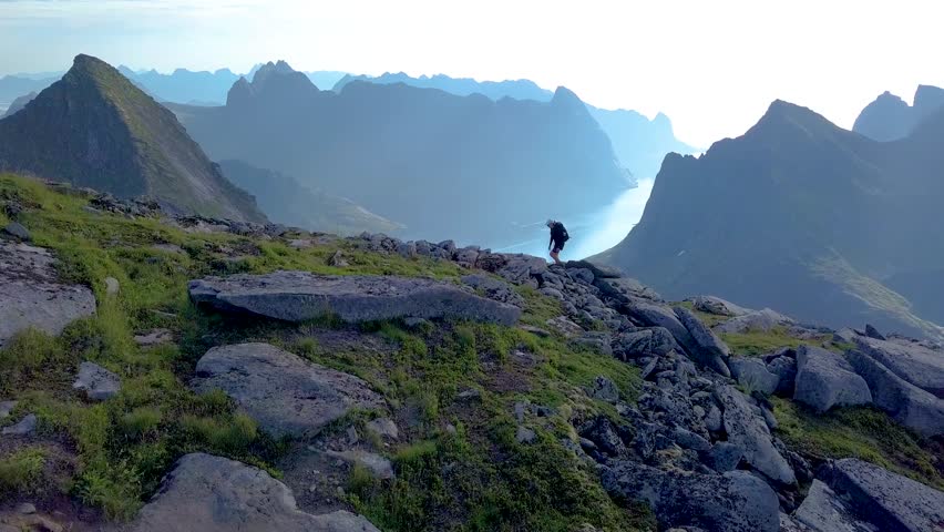 Aerial view of girl with a backpack rises on a mountain ridge. Beautiful view of the peaked tops of the Lofoten Islands. Norway 4k | Shutterstock HD Video #1015725367