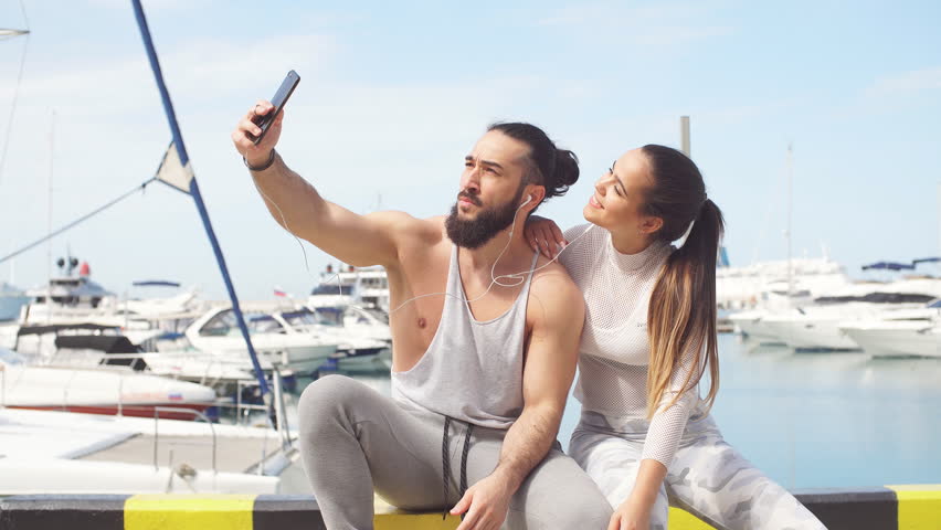 Outdoor view of loving young athletic couple in sportswear on summer vacation taking a self portrait with smartphone on seafront