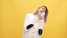 Cheerful woman in warm cardigan and headphones listening music by smartphone and sings over yellow background