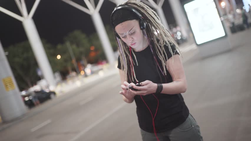 Young attractive woman with dreadlocks use smartphone listening music on headphones. On street at night 