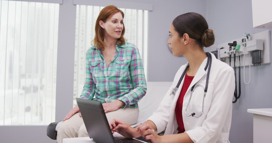 Portait of young latina doctor using laptop computer while talking with senior patient inside clinic. Close up of middle aged patient discussing with young female doctor health issues Royalty-Free Stock Footage #1015761151