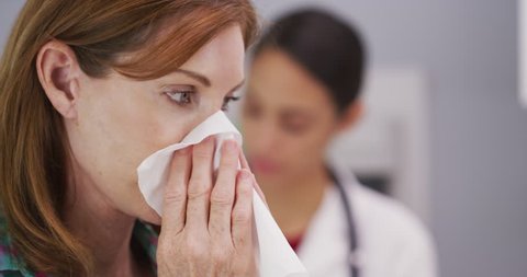 Attractive mid aaged patient blowing nose into tissue paper. Close up of caucasian woman wiping nose with tissue while doctor sits quietly in the background taking notes