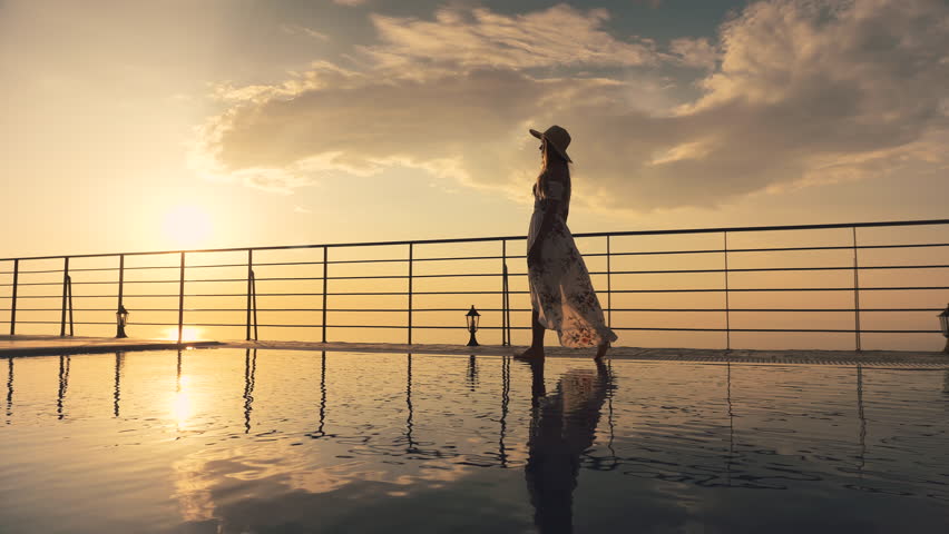 Slow motion - Beautiful shot of a woman in a long flow dress walking by the poolside and splashing water with her foot at colorful sunset. Girl leaning on the railing to enjoy the view at end of clip Royalty-Free Stock Footage #1015761727