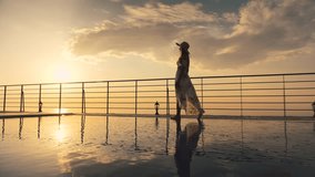Slow motion - Beautiful shot of a woman in a long flow dress walking by the poolside and splashing water with her foot at colorful sunset. Girl leaning on the railing to enjoy the view at end of clip