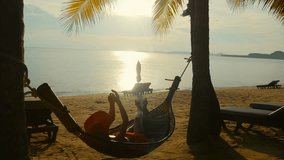 Young happy lady in orange hat relaxing in the hammock on the sandy beach and enjoying sunset. The concept of a winter holiday on the tropical island.