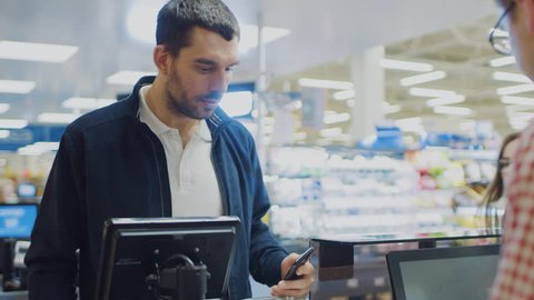At the Supermarket: Checkout Counter Customer Pays with Smartphone for His Food Items. Big Shopping Mall with Friendly Cashier, Small Lines and Modern Wireless Paying Terminal System.