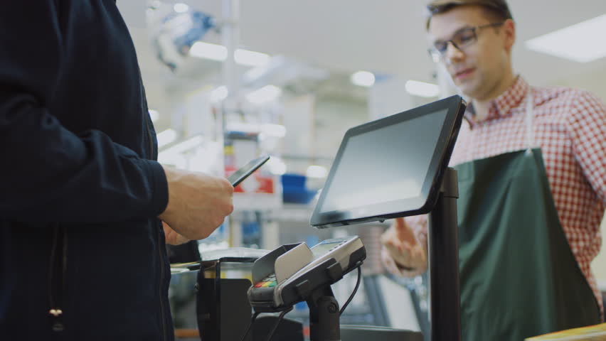 At the Supermarket: Checkout Counter Customer Pays with Smartphone for His Food Items. Big Shopping Mall with Friendly Cashier, Small Lines and Modern Wireless Paying Terminal System. Royalty-Free Stock Footage #1015778251