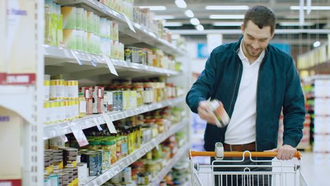 At the Supermarket: Handsome Man Browses Through Shelf with Canned Goods, Chooses Tin Can and Places it into His Shopping Cart. Shot on RED EPIC-W 8K Helium Cinema Camera.
