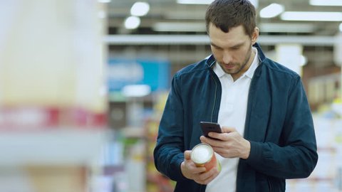 At the Supermarket: Handsome Man Uses Smartphone to Check Nutritional Value of the Canned Goods and Buy it. He's Standing with Shopping Cart in Canned Goods Section. Shot on RED EPIC-W 8K Camera.