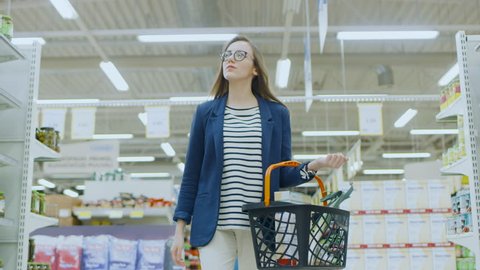 At the Supermarket: Beautiful Young woman with Shopping Basket Walks Through Canned Goods Section, Browsing. Big Store with Lots of Aisles. Shot on RED EPIC-W 8K Helium Cinema Camera.