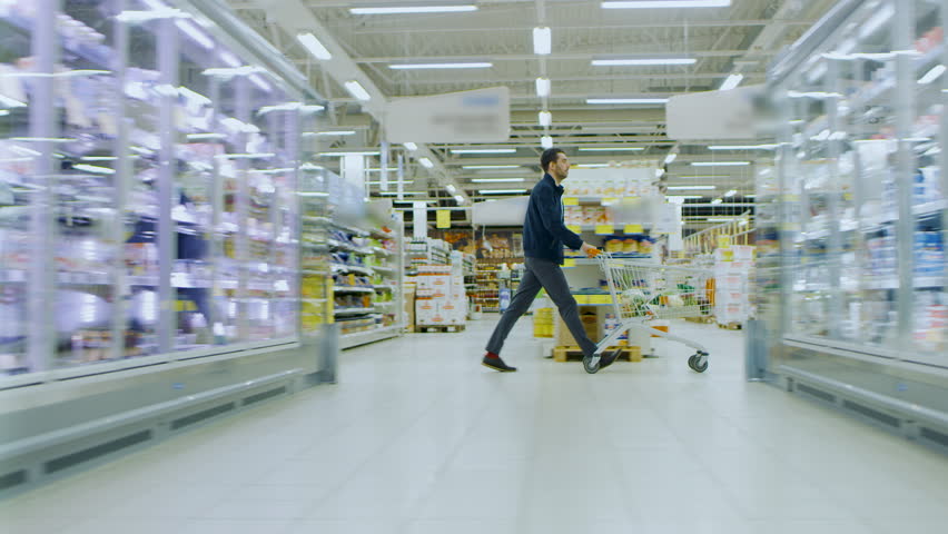 At the Supermarket: Man in a Hurry Pushes Shopping Cart full of Items, He's Walking Through Different Section of the Big Bright Mall. Following / Moving Side view Footage. Shot on RED EPIC-W 8K. Royalty-Free Stock Footage #1015782340