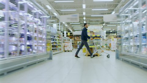 At the Supermarket: Man in a Hurry Pushes Shopping Cart full of Items, He's Walking Through Different Section of the Big Bright Mall. Following / Moving Side view Footage. Shot on RED EPIC-W 8K.