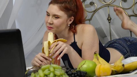 Young woman enjoying on a bed, using laptop and eating a banana at home in the morning. slow motion