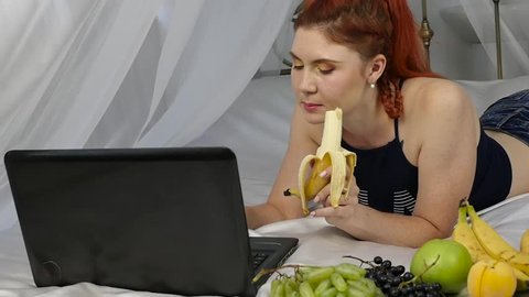Young woman enjoying on a bed, using laptop and eating a banana at home in the morning. slow motion