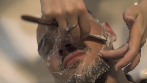 Barber shaving bearded man with straight razor in male salon. Close up shaving razor to hipster beard in barber shop. Male skin care and beard style concept