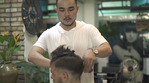 Asian hairdresser doing male haircut with electric shaver. Man getting haircut in barber shop. Professional hairstylist cutting hair in male salon. Man hairdressing with electric razor