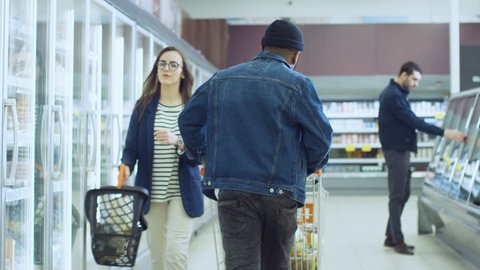 At the Supermarket: Happy Stylish Guy Pushes Shopping Cart and Chooses Products in the Frozen Goods Section of the Store. Big Mall with Glass Door Fridge. Shot on RED EPIC-W 8K Helium Cinema Camera.