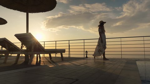 Beautiful shot of a woman in a long flow dress walking by the poolside and splashing water with her foot at colorful sunset Video Stok