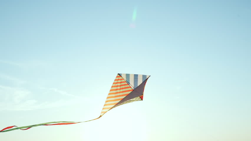 Bright kite with long tail of silk ribbons flies in light wind against blue sky in rays of sun at sunset of day slow motion. Symbol of Liberty and Childhood. Freedom