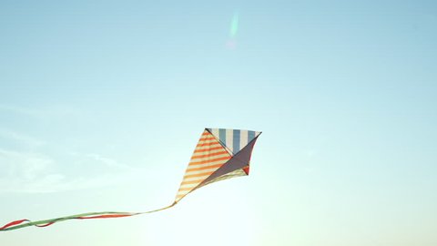 A slow motion bright kite with a long tail of silk ribbons flies in a light wind against the blue sky in the rays of the sun at sunset of the day. The Symbol of Liberty and Childhood