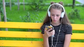 Teenage girl with headphones talking while looking at smart phone and sitting down on bright yellow bench