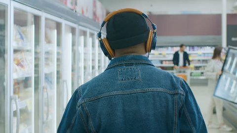 At the Supermarket: Stylish African American Guy with Headphones Walks Through Frozen Goods Section of the Store. Following Back View Shot. Slow Motion.