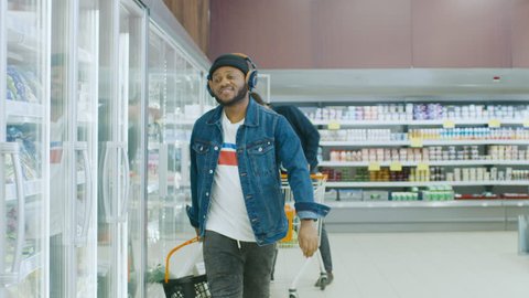 At the Supermarket: Stylish African American Guy with Shopping Basket Chooses Products in the Frozen Goods Section of the Store. He Opens Big Glass Door Fridge. Slow Motion. Shot on RED EPIC-W 8K.