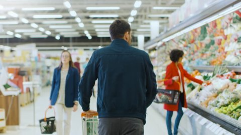 At the Supermarket: Man Pushing Shopping Cart Through Fresh Produce Section of the Store. Store with Many Customers Shopping. Following Back View Shot. Shot on RED EPIC-W 8K Helium Cinema Camera.