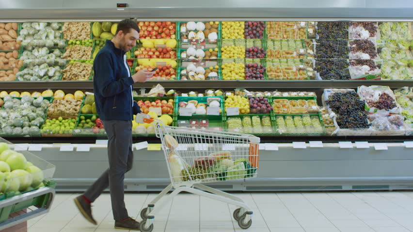 At the Supermarket: Handsome Man with Smartphone, Pushes Shopping Cart, Walks Past Fresh Produce Section of the Store. Man Immersed in Internet Surfing on His Mobile Phone. Side View Shot.  Royalty-Free Stock Footage #1015805194