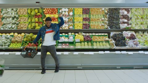 At the Supermarket: Happy Stylish Guy with Shopping Basket Dances Through Fresh Produce Section of the Store. Big Bright Shopping Mall with Lots of Goods and Products. Shot on RED EPIC-W 8K Camera.