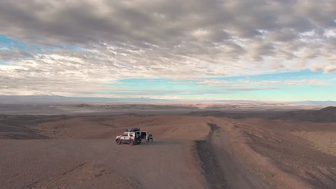 Aerial cinematic view of two people and car at road trip in sand desert Atacama Chile. People sitting and watching landscape at sunrise. Drone rising up filming forward. Video de stock