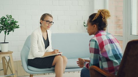 Confident female psychologist is holding consultation with African American girl, listening to her and writing while patient is speaking explaining problem.