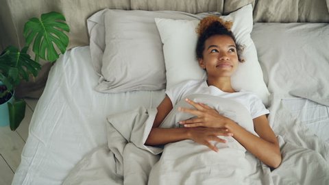 Happy mixed race girl is lying in bed and smiling then stretching arms, closing eyes and falling asleep. Resting people, bedtime and interiors concept.