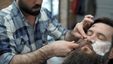 Barber shaves men with a long beard with straight razor blade in s hair salon or barbershop. Man's haircut and shaving at the hairdresser, barber shop and shaving salon.