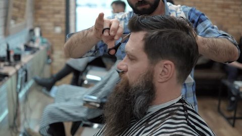 Close-up of barber cuts the hair by scissors at barbershop. Hairdresser's hands at working process. Barber making haircut of attractive bearded man in barbershop. Hairdresser at work. Beauty saloon.
