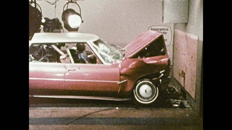1980s: Car slams into barricade on vehicle test track. Crash test dummies lie in crashed car. Man talks and stands near dolly cart with crash test dummies.